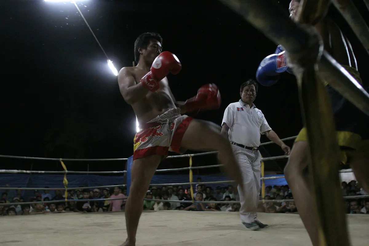 A Kun Khmer fighter in red gloves and shorts lifts his knee in a floodlit ring while a referee in white watches and a crowd stands behind the ropes.