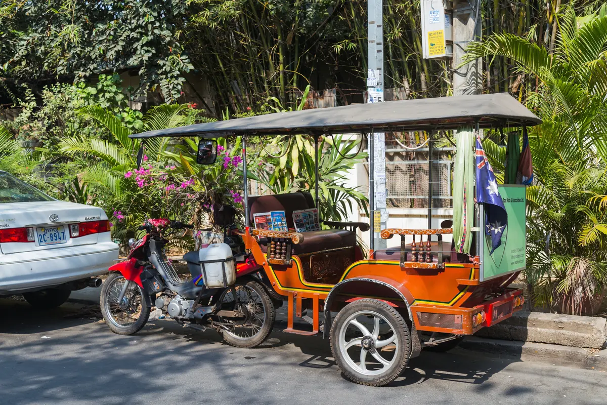A parked Cambodian tuk-tuk, a motorbike pulling a covered two-bench carriage, in front of green palms on a Phnom Penh street.