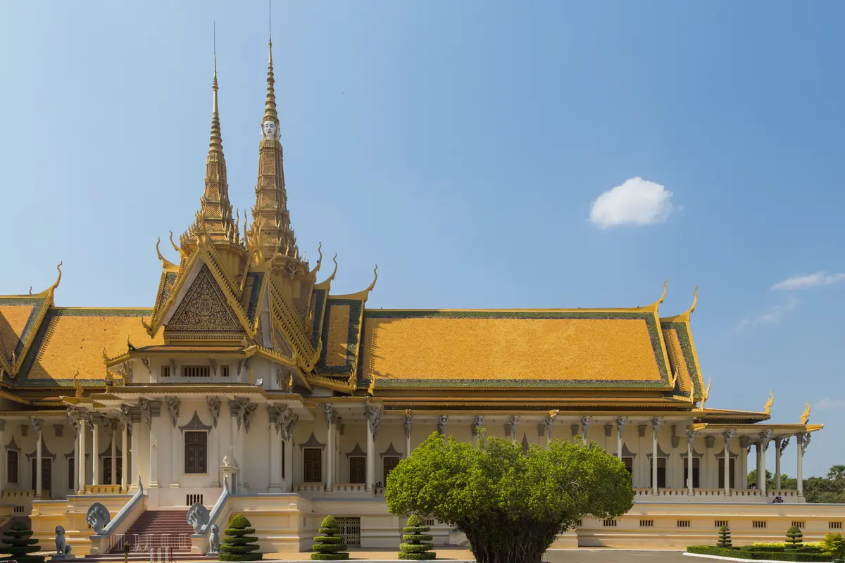 The golden-roofed Throne Hall of the Royal Palace in Phnom Penh, with two tall spires, white columns and a banyan tree in front under a blue sky