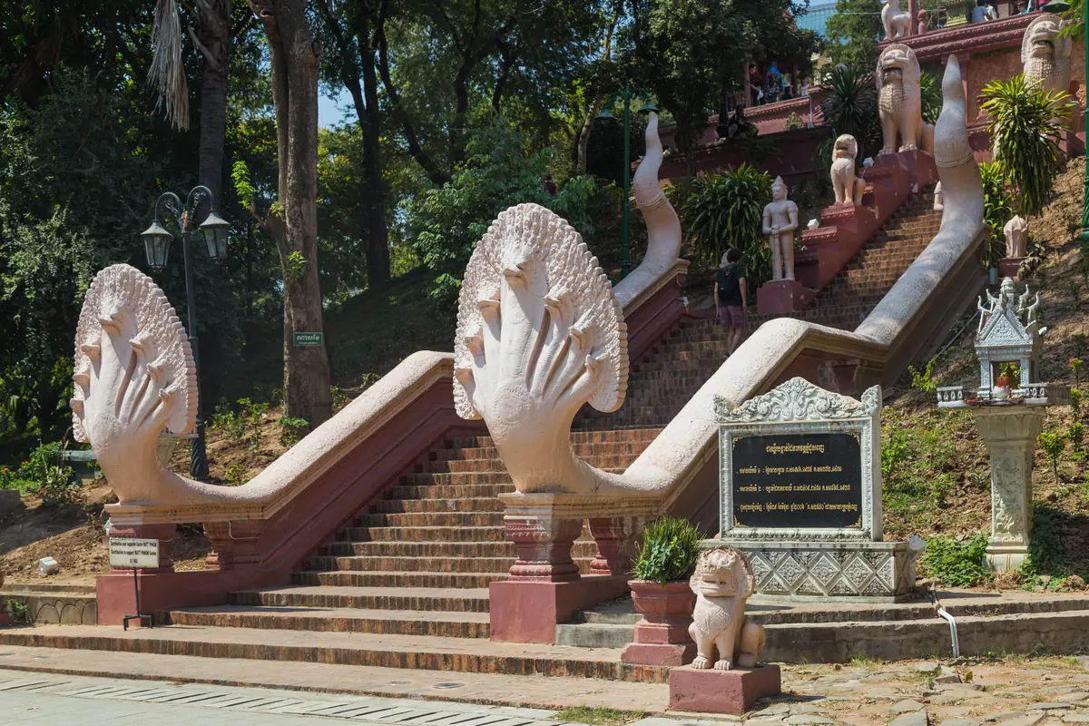 A broad stairway of brick steps flanked by white and pink multi-headed naga balustrades and stone lions climbing a wooded hill to Wat Phnom